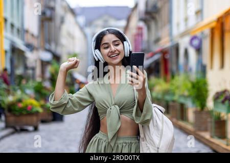 Jeune femme belle ID marche dans la ville, touriste tient le téléphone dans les mains, utilise des écouteurs pour écouter de la musique et des podcasts de radio en ligne, danse et chante joyeusement. Banque D'Images