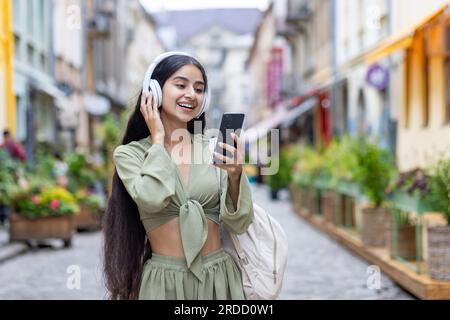 Jeune femme belle ID marche dans la ville, touriste tient le téléphone dans les mains, utilise des écouteurs pour écouter de la musique et des podcasts de radio en ligne, danse et chante joyeusement. Banque D'Images