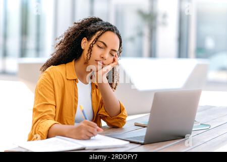 Dormir au travail ou étudier. Fille brésilienne ou latino aux cheveux frisés fatiguée, assise à l'extérieur avec un ordinateur portable, endormie en étudiant ou en travaillant en ligne, se sentant fatiguée et manque de sommeil, sieste les yeux fermés Banque D'Images