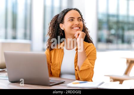 Fille aux cheveux bouclés brésilienne ou hispanique positive, freelance, étudiante, spécialiste DE L'INFORMATIQUE, assis à l'extérieur, avec ordinateur portable, semble heureux sur le côté, pensant à quelque chose de bon, rêvant, souriant Banque D'Images