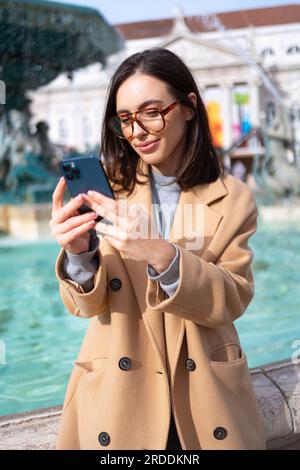 Femme portant des lunettes, assise à la fontaine, tenant le smartphone et souriant doucement. Bonnes vibrations et sentiments. Communication en ligne à l'aide d'un appareil numérique mobile Banque D'Images