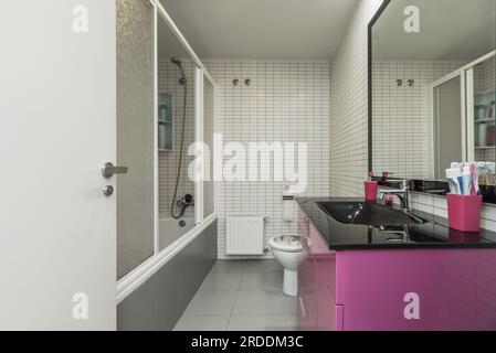 Une salle de bains avec un lavabo noir brillant, armoire en bois avec tiroirs violets profonds, baignoire avec écrans, miroir encadré noir et carreaux de métro blancs Banque D'Images