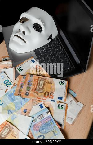 Sur un ordinateur portable, il y a un masque blanc et une pile de billets en euros, concept de cybercriminalité Banque D'Images