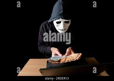 Hacker homme habillé en noir avec masque blanc et capuche noire comptant les billets en euros dans ses mains, concept de cybercriminalité, isolé sur fond noir Banque D'Images