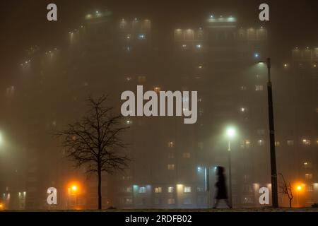 Soirée effrayante avec un brouillard dense couvrant la rue avec un bâtiment résidentiel et des lanternes brillantes avec la silhouette de la personne qui marche Banque D'Images