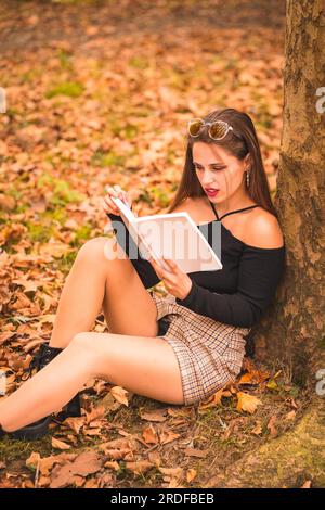 Portrait d'une femme en automne dans une forêt avec des feuilles brunes assise lisant un livre Banque D'Images