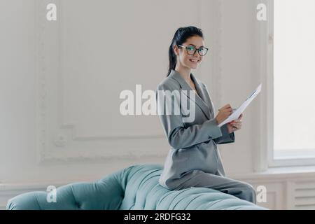 Femme prospère gestionnaire financier étudie les documents dans un bureau à domicile spacieux, habillé avec élégance, écrivant des informations, regarde heureux à la caméra. PAP Banque D'Images