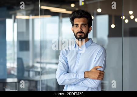 Portrait d'un jeune programmeur indien réussi, développeur debout dans un centre de bureau, croisant ses bras sur sa poitrine et regardant sérieusement la caméra. Banque D'Images