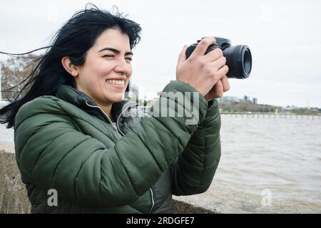 Portrait de jeune femme latine vénézuélienne touriste souriant dehors heureux de faire une photo avec son appareil photo numérique DSLR, debout sur la jetée par la riv Banque D'Images