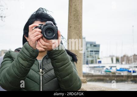 Vue de face de la jeune femme debout sur la jetée au bord de la rivière à l'aide d'un appareil photo numérique DSLR créant du contenu pour les médias sociaux. Banque D'Images