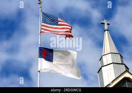 American flag and Christian flag fly besides a church steeple.  Cross seems to stand between or besides the two. Stock Photo