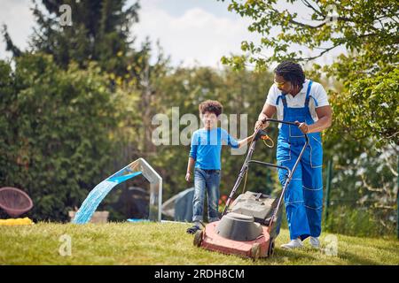 Jeune père afro-américain tondant de l'herbe à l'extérieur avec son jeune fils. Travailler ensemble dans l'arrière-cour. Famille, unité, concept de style de vie. Banque D'Images