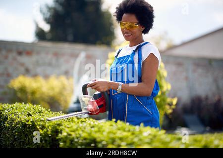 Femme afro-américaine alors qu'elle tond habilement des haies à l'aide d'un taille-haie. Avec un comportement confiant et concentré, elle façonne habilement le feuillage, crea Banque D'Images