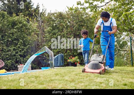 Jeune père tondant de l'herbe avec une tondeuse à gazon électrique à l'extérieur tandis que son jeune fils l'aide. Travailler ensemble en équipe, père et fils. Accueil, lif Banque D'Images