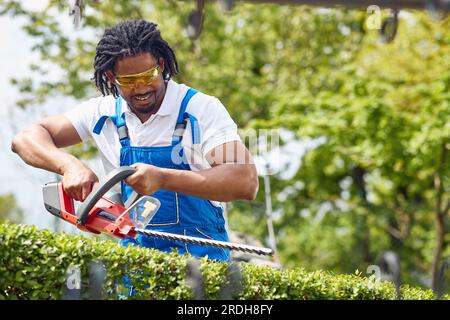 Jeune homme afro-américain en costume de travail à l'aide d'un coupe-herbe électrique pour couper la haie en plein air, jardinage. Maison, style de vie, concept de jardinage. Banque D'Images