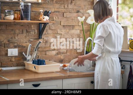 Femme avec un sourire radieux comme elle se tient dans sa cuisine. Avec ses mains immergées dans l'eau savonneuse et un comportement concentré mais détendu, la femme s'efforce Banque D'Images