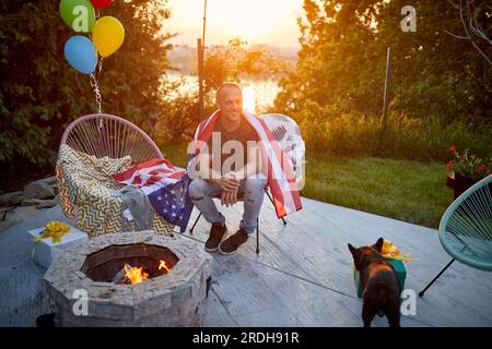 Joyeux homme d'âge moyen avec drapeau américain autour de son dos, assis à l'extérieur près d'une cheminée dans son jardin, profitant d'une journée d'été ensoleillée avec son chien. Li Banque D'Images