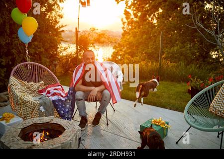 Joyeux jeune homme assis à l'extérieur dans le jardin par une cheminée se sentant heureux avec ses deux chiens qui courent autour de la cour arrière. Lifestyle, lesiure, 4e. Banque D'Images