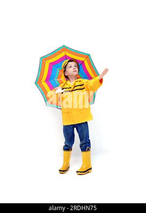 Young boy in yellow raincoat and rubber boots holds his hand out to check for rain.  He is holding a colorful striped umbrella. Stock Photo