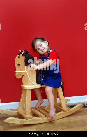 Petite fille monte son cheval à bascule en bois et rêve d'avoir un jour un vrai cheval. Les murs sont rouge foncé et la petite fille porte une chemise rouge et d Banque D'Images