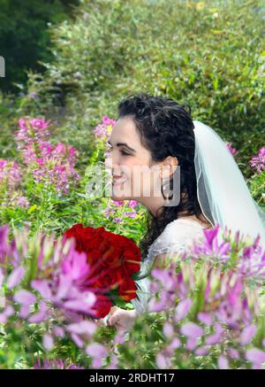 Mariée, tenant des roses rouges, est entourée de fleurs roses dans un jardin extérieur. Elle sourit et rit et la joie rayonne de son visage. Banque D'Images