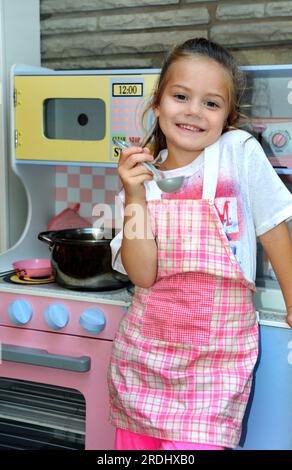 Une petite fille « joue des actes » étant une maman et cuisinant pour sa famille. Elle porte un tablier à motif écossais rose et un sourire. Elle tient également une cuillère de dégustation Banque D'Images