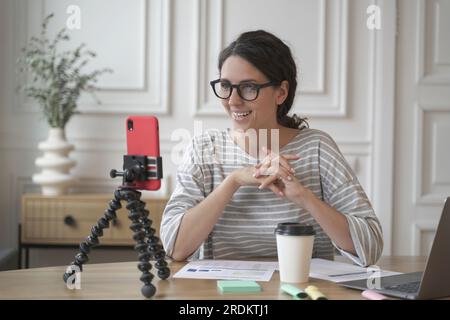 Jeune femme d'affaires espagnole souriante dans des lunettes assis devant un smartphone sur un trépied sur le lieu de travail, influenceur positif femelle enregistrant la vidéo pour b Banque D'Images