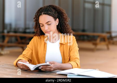 Calme concentré brésilien ou hispanique étudiant aux cheveux bouclés, assis à l'extérieur avec un livre dans ses mains, la lecture concentrée d'un livre intéressant, obtenir des informations, s'inspirer. Concept de lecture Banque D'Images