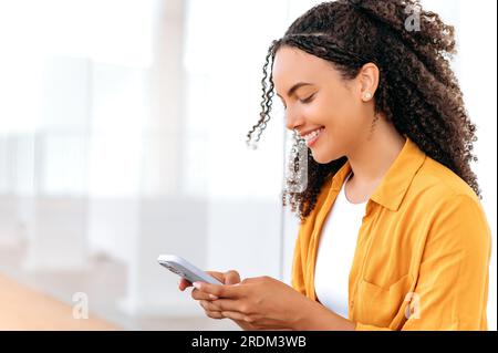 Utilisation de gadgets. Vue de côté gros plan photo d'une jolie femme hispanique ou brésilienne aux cheveux bouclés, utilise son téléphone intelligent, lit des nouvelles, messagerie dans les médias sociaux, texto avec la famille, sourire heureux Banque D'Images