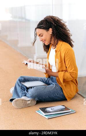 Photo verticale de belle étudiante brésilienne ou hispanique aux cheveux bouclés positive et élégante, asseyez-vous près du campus universitaire, lisez un livre, préparez-vous à l'examen, faites vos devoirs, inspirez-vous, souriez Banque D'Images