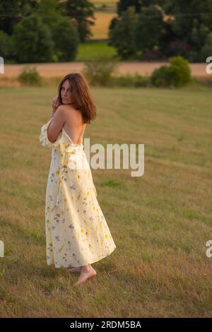 Une jeune belle dame marche pieds nus dans un champ au coucher du soleil. Une fille dans une belle robe jaune pose au coucher du soleil dans un champ. Banque D'Images