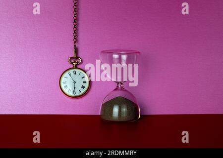 Minuterie de sable en verre et montre de poche dorée suspendue sur un fond rose et une base rouge Banque D'Images