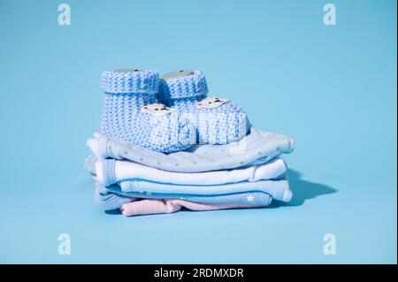 Natures mortes bottes de bébé tricotées bleues sur une pile de bodys nouveau-nés repassés lavés propres, isolés sur fond pastel bleu. Préparations du sac Banque D'Images