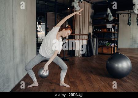 Femme aux cheveux roux dans la salle de gym, faisant plie avec fitball à la main, embrasse l'ambiance sportive. Activités de remise en forme présentées. Banque D'Images