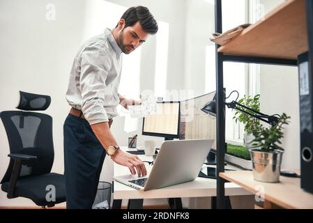 Multitâche. Vue de côté du commerçant barbu réussi ou homme d'affaires en tenue formelle travaillant avec un ordinateur portable tout en se tenant debout dans son bureau moderne. Banque D'Images