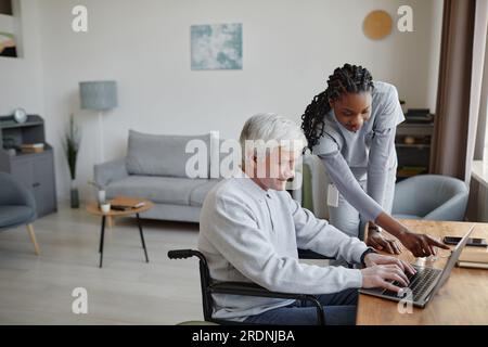 Portrait vue de côté de la jeune femme noire comme infirmière à domicile aidant l'homme âgé en fauteuil roulant à l'aide de l'ordinateur, espace de copie Banque D'Images
