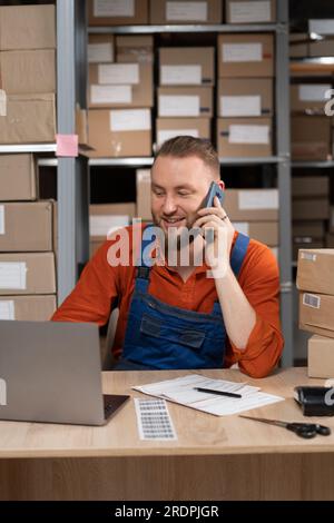 Gestionnaire d'entrepôt parlant au téléphone tout en souriant assis avec l'ordinateur portable à la table Banque D'Images