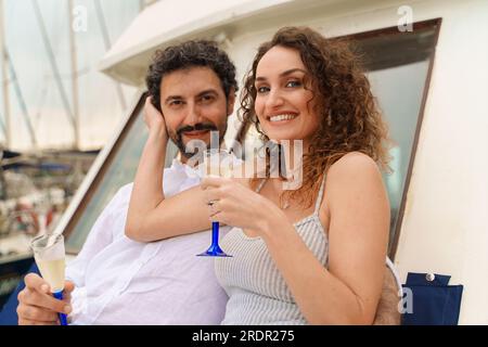 Un jeune couple de jeunes mariés, un homme caucasien brunette avec une barbe et une brésilienne à la peau claire avec les cheveux bouclés, appréciant du champagne sur un pont de bateau. Banque D'Images