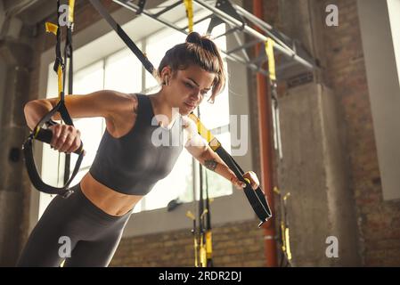 Push-up parfait. Vue latérale de jeune femme sportive avec un corps parfait en vêtements de sport faisant des pompes avec des sangles de fitness trx dans la salle de gym. Banque D'Images