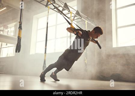 Push-up parfait. Vue latérale d'un homme sportif jeune et fort en vêtements de sport faisant des pompes avec des sangles de fitness trx à la salle de gym Banque D'Images