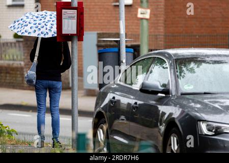 Duisburg, Allemagne. 23 juillet 2023. Une femme se tient debout avec un parapluie à un arrêt de bus à Duisburg. Les habitants de Rhénanie-du-Nord-Westphalie doivent se préparer aux jours de pluie. Crédit : Christoph Reichwein/dpa/Alamy Live News Banque D'Images