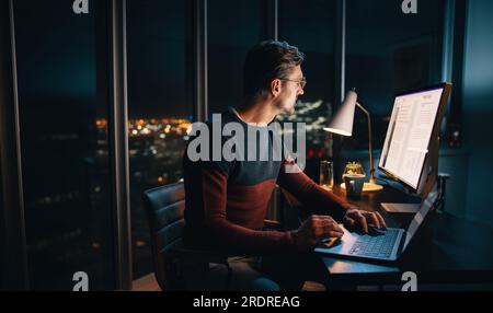 Un homme d'affaires concentré travaille tard dans un bureau bien éclairé, tapant sur son ordinateur portable. Entouré d’un milieu de travail occupé, il se concentre avec diligence sur un plan de marketing Banque D'Images