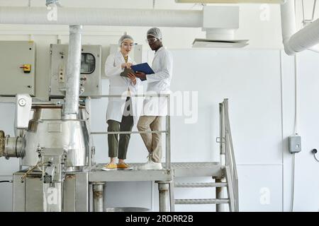 Portrait en pied de deux travailleurs portant des blouses de laboratoire tout en supervisant la production dans l'atelier d'usine, espace de copie Banque D'Images
