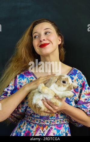 Belle femme russe aux longs cheveux blonds tenant un animal de compagnie de lapin dans ses mains contre le mur noir Banque D'Images