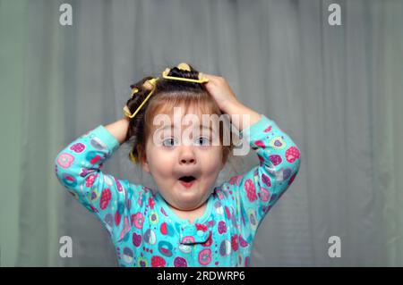 Les bigoudis éponge à l'ancienne donnent envie à cette petite fille de les sortir. Elle fait un visage comique en mettant ses mains sur sa tête. Banque D'Images