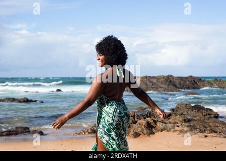 Portrait de belle femme aux cheveux noirs souriante et heureuse debout sur la plage. En arrière-plan, le ciel cloue les rochers et la mer. Rio Vermelho, Salvad Banque D'Images