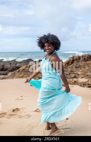 Belle femme avec les cheveux de puissance noirs portant la tenue longue bleu clair dansant sur le sable de plage. Femme heureuse et confiante. Ciel et nuages dans le backg Banque D'Images