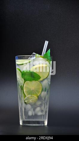 Un grand verre à facettes d'un mélange de jus d'agrumes avec de la glace, des pailles et des tranches de citron et de citron vert sur un fond noir. Disposition verticale. Banque D'Images