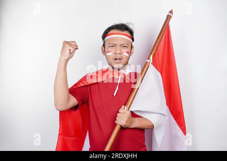 Un jeune homme asiatique avec une expression heureuse et réussie portant un haut rouge et un bandeau tout en tenant le drapeau indonésien, isolé par fond blanc. Indone Banque D'Images