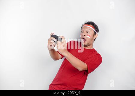 Un portrait d'un homme asiatique choqué portant un bandeau, tenant son téléphone alors que sa bouche grande ouverte, isolé par fond blanc. L'indépendance de l'Indonésie Banque D'Images